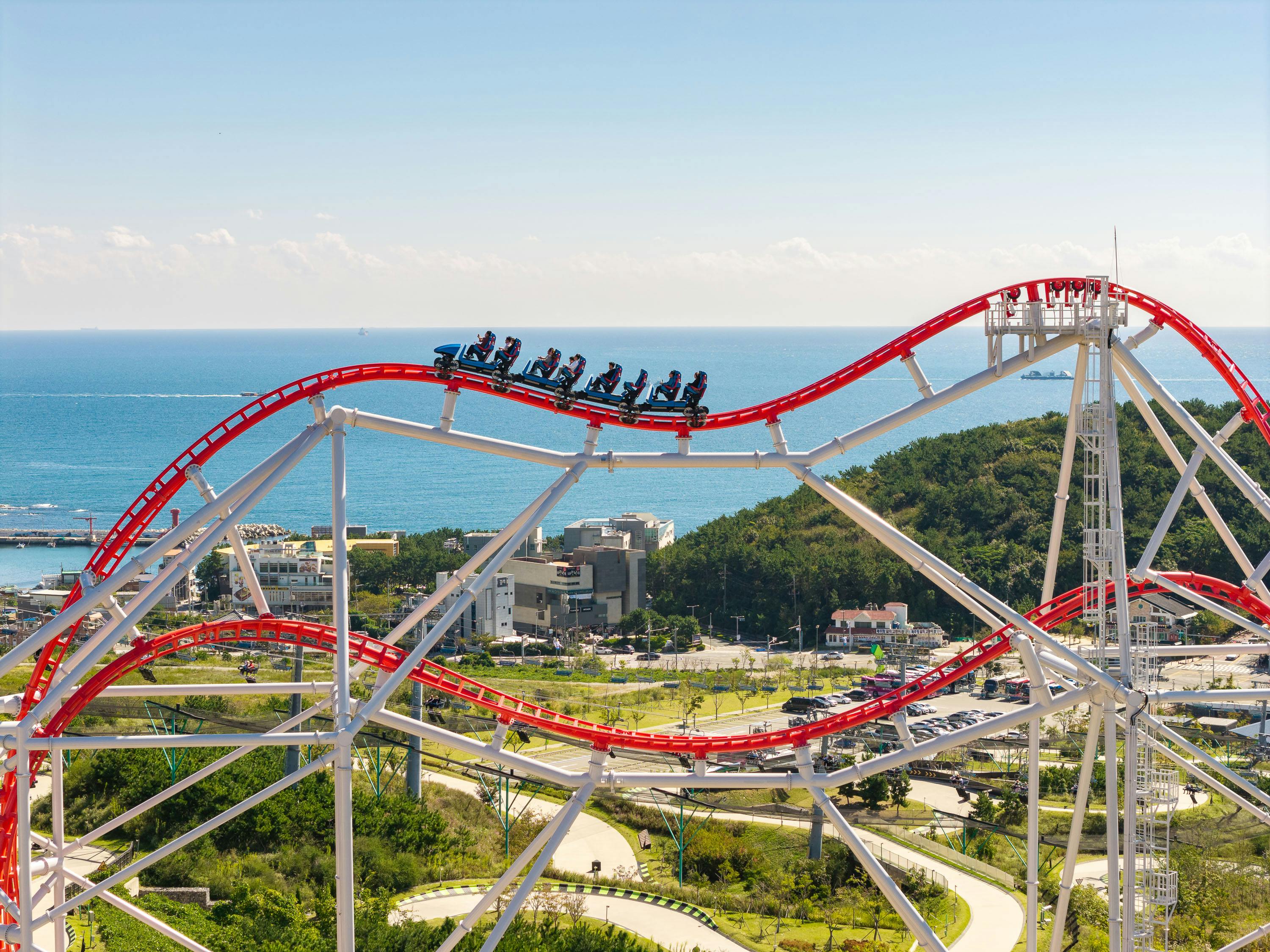 A red roller coaster with passengers riding through loops, set against a seaside landscape and clear blue sky.