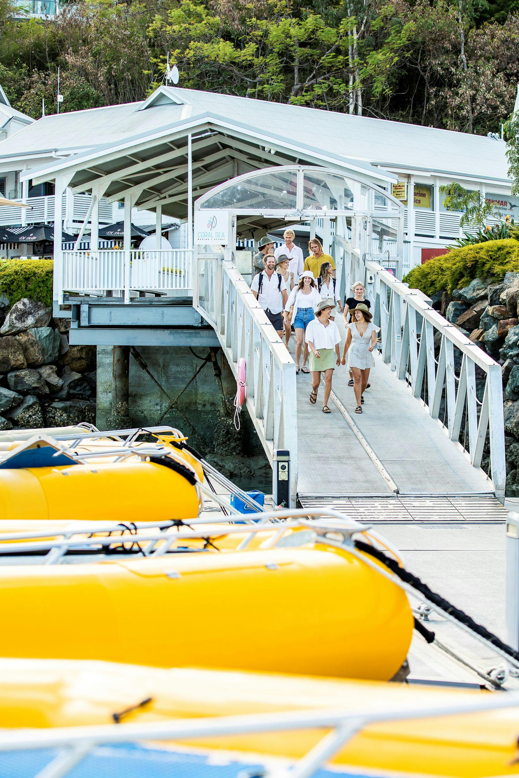 A group of people walking down a dock ramp towards yellow boats, with a white building and greenery in the background.