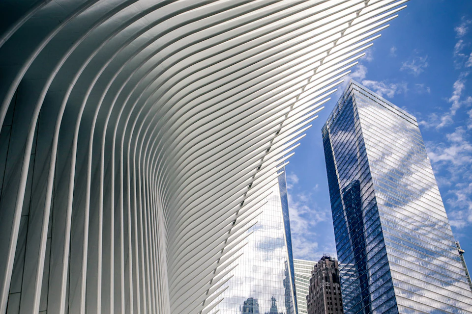 Curved, white architectural ribs on the left frame modern glass skyscrapers against a blue sky with clouds.