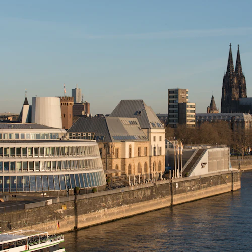 En modern, cirkulär byggnad och historiska strukturer längs en flod, med en stor katedral med tvillingspiror i bakgrunden.