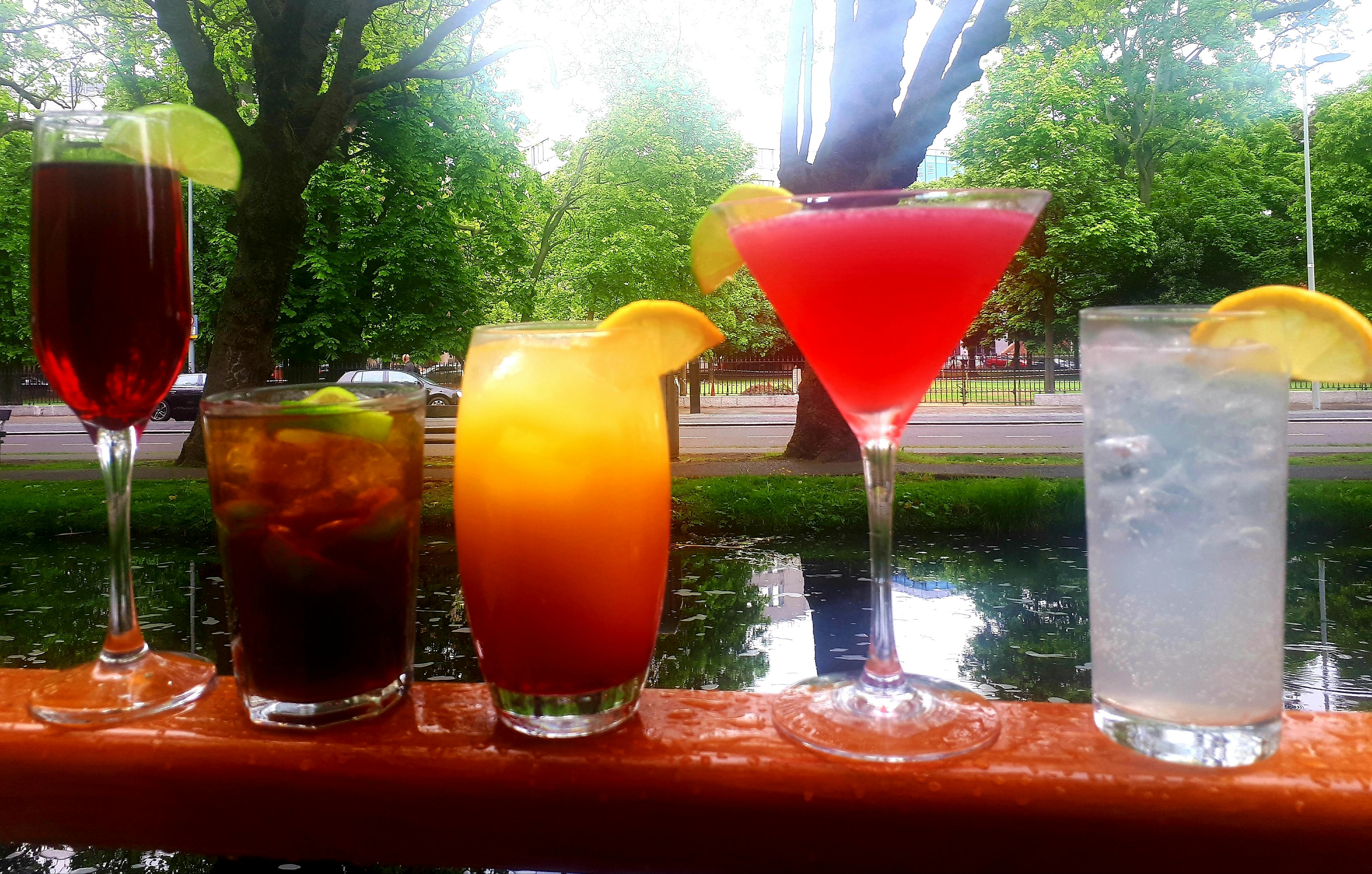 四种五颜六色的鸡尾酒，包括一杯红马提尼，陈列在栏杆上，背景是绿树成荫的公园。