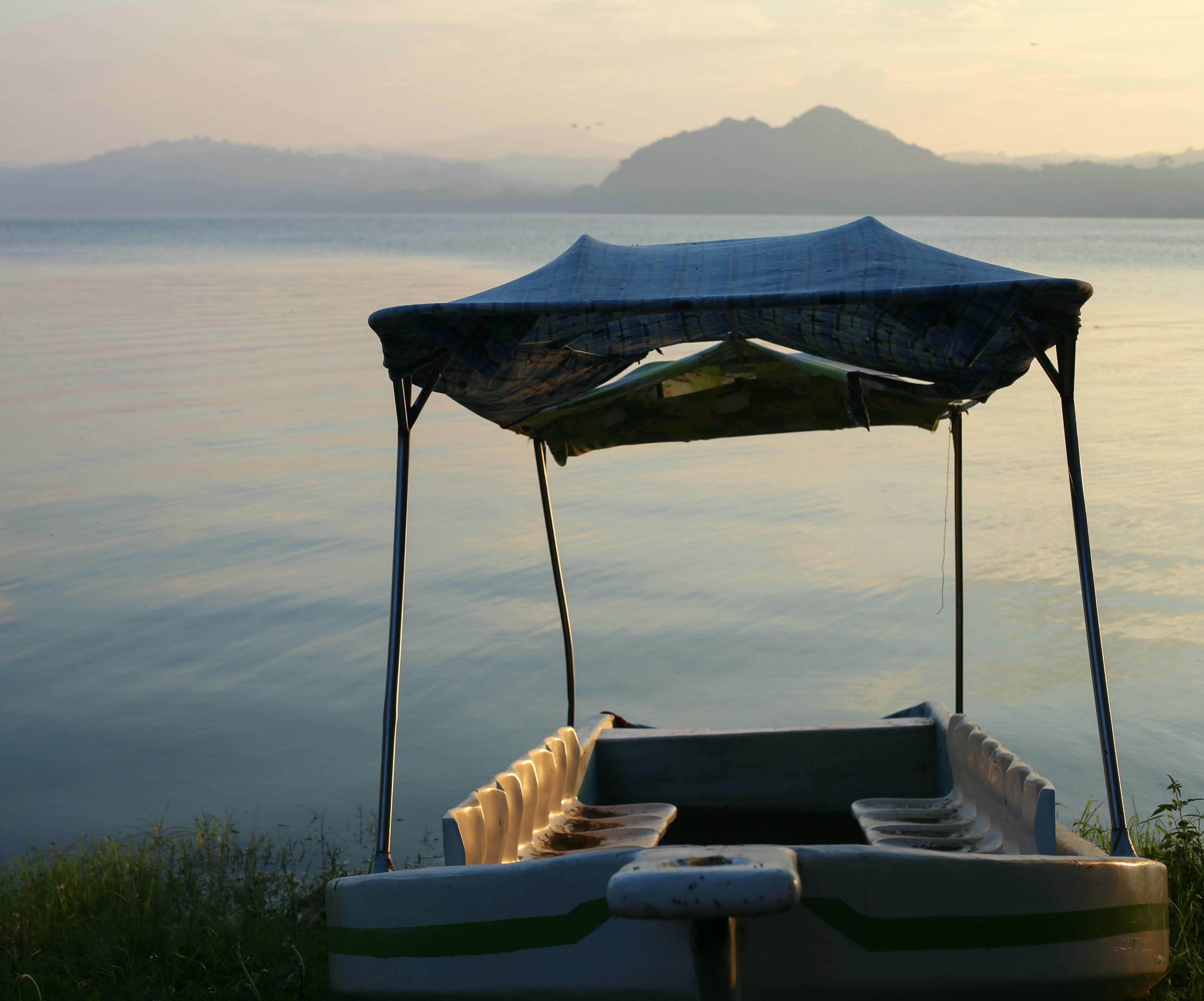 Prázdná loď s baldachýnem na klidné vodě u břehu; kopce jsou při západu slunce slabě viditelné v pozadí.
