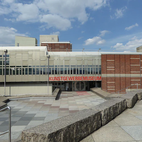 En modern byggnad med rött tegel och glas exteriör, med "Kunstgewerbemuseum" skrivet i rött ovanför ingången.