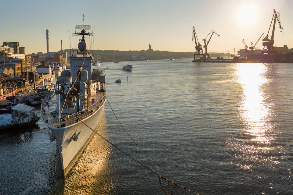 A docked naval ship with a radar tower against a sunset, cranes, and other ships in the harbor.