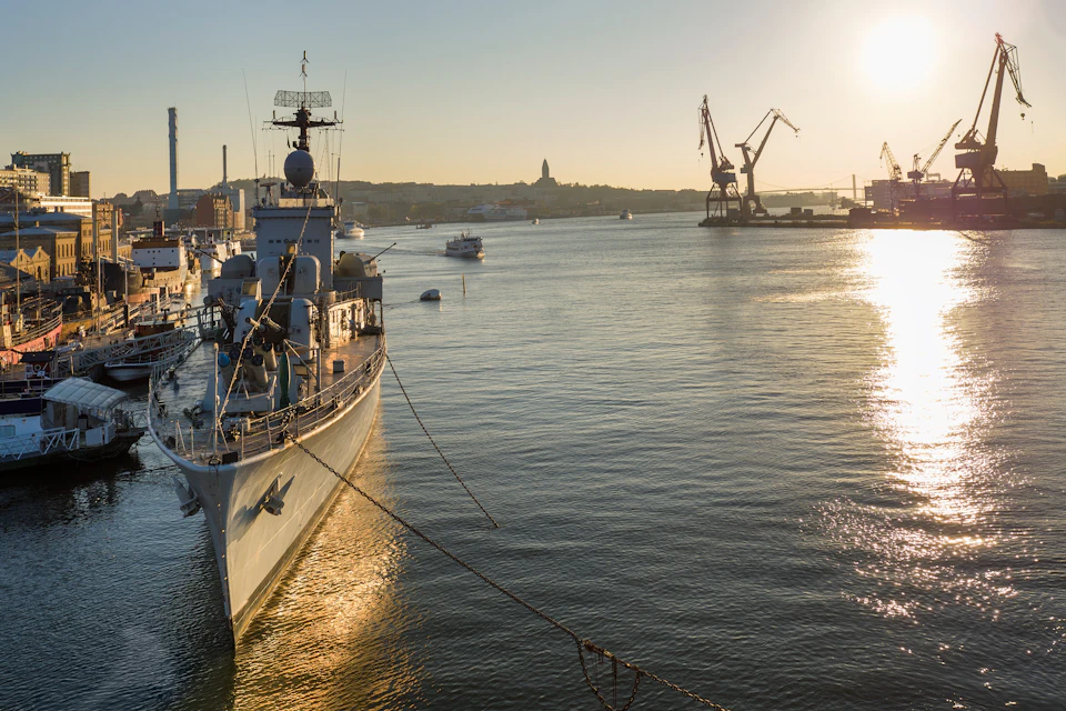A docked naval ship with a radar tower against a sunset, cranes, and other ships in the harbor.