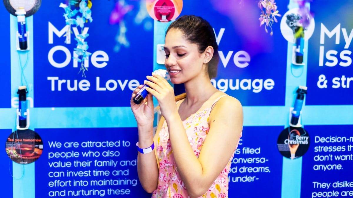 A woman in a pink floral dress holds a microphone, speaking in front of a blue backdrop with text.