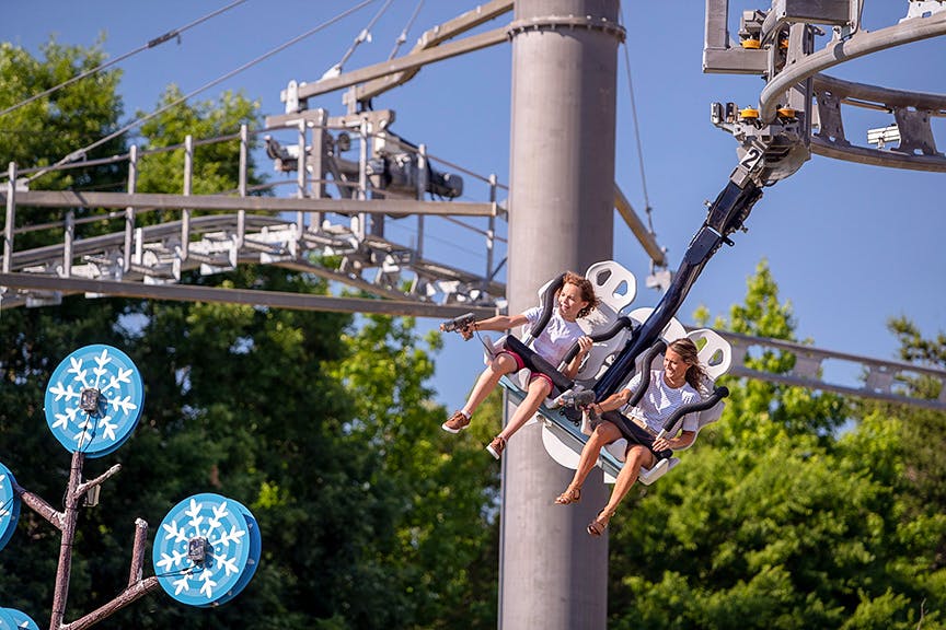 Two people are seated on a suspended amusement ride, smiling and enjoying the experience. Trees and a clear blue sky are in the background.