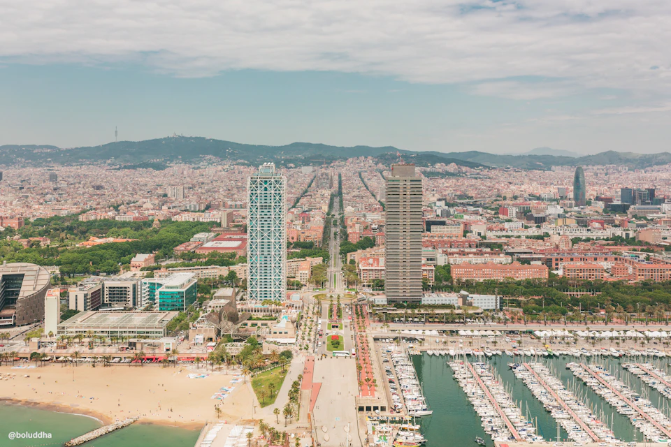 Aerial view of Barcelona's coastline featuring two tall buildings, a marina with boats, and sandy beaches with people.