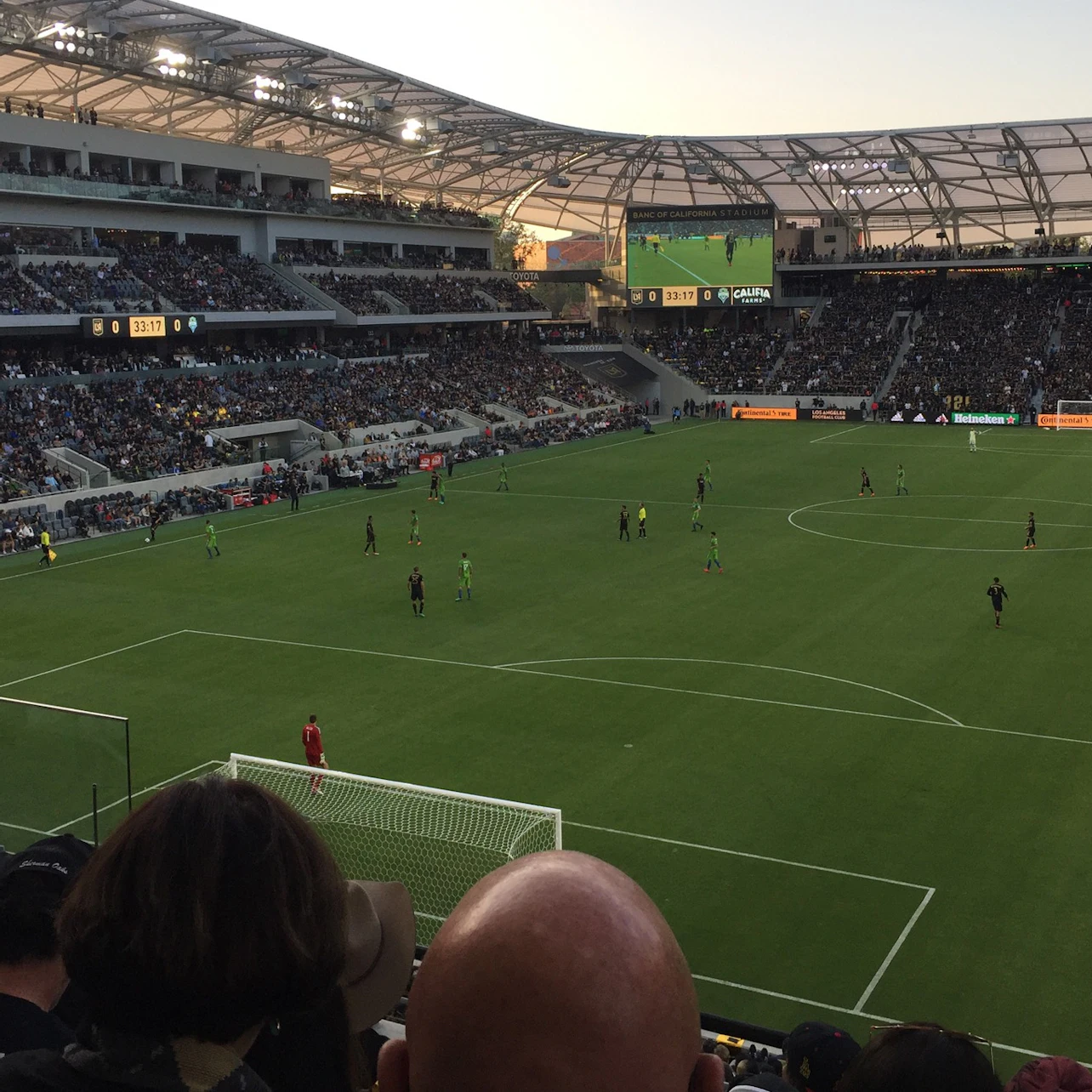 A soccer stadium filled with spectators, a match in progress on a green field under a partially covered roof.