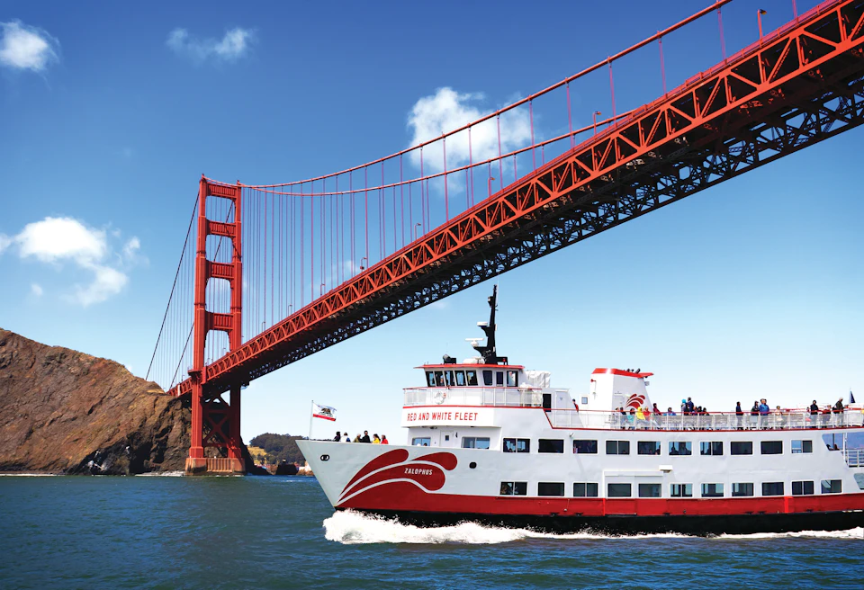 A boat labeled "RED AND WHITE FLEET" passes under the Golden Gate Bridge on a sunny day.