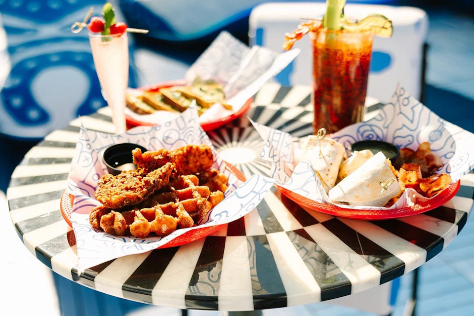 A table with plates of waffles, fried chicken, sandwiches, and drinks, including a Bloody Mary and a blended drink.