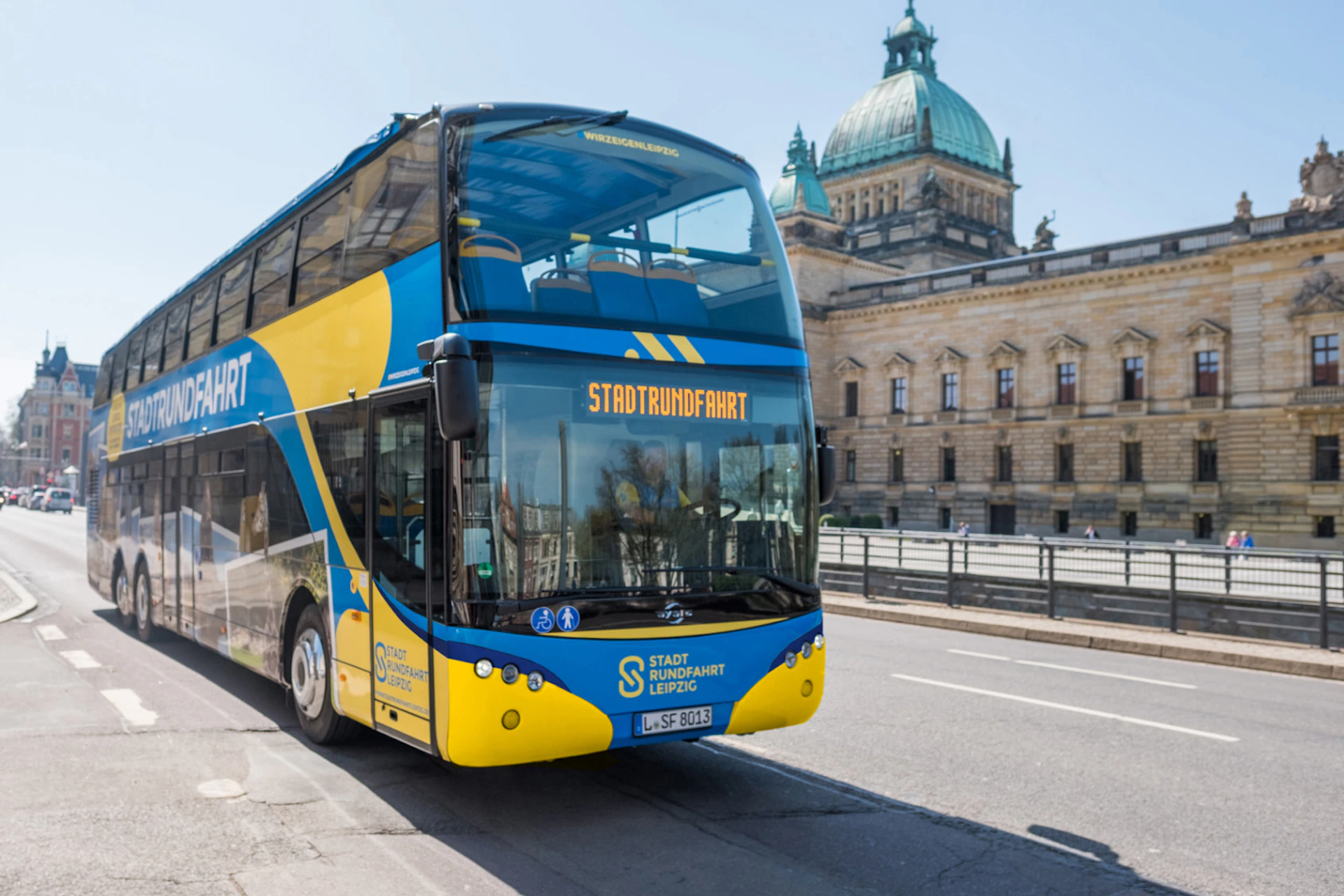A yellow and blue double-decker tour bus in front of a historic building with a dome and statues, under a clear blue sky.