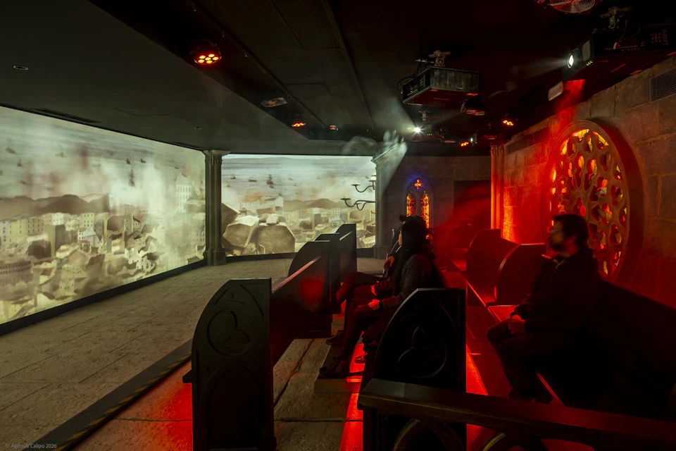 People seated in a dark immersive theatre with atmospheric lighting and large surrounding projection screens displaying a fiery cityscape.