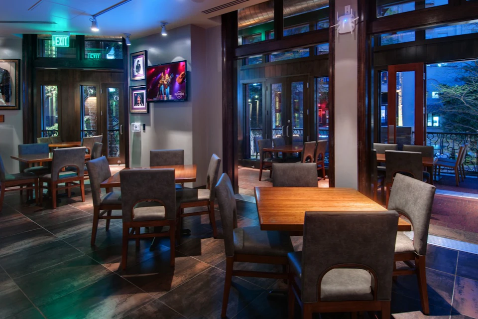 A modern restaurant interior with wooden tables, upholstered chairs, and a television displaying a music performance.