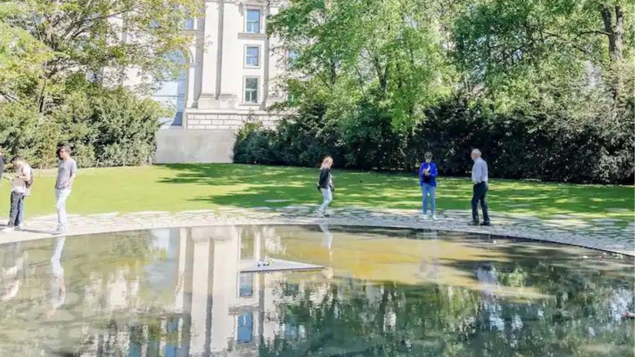 Tři lidé stojí na trávníku u reflexního rybníka v parku s budovou a stromy v pozadí.