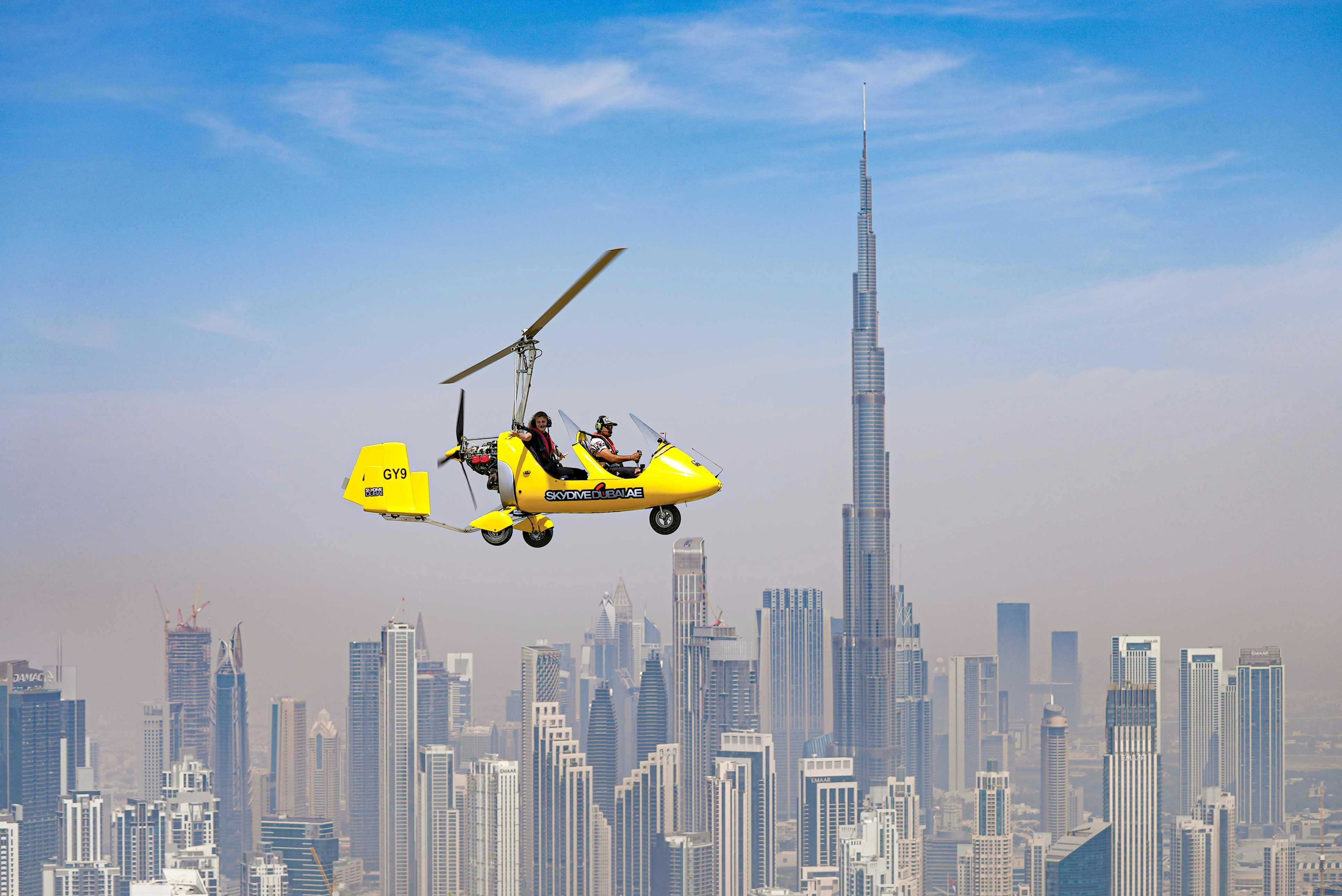 Um girocóptero amarelo a sobrevoar uma paisagem urbana com arranha-céus modernos, incluindo o Burj Khalifa em segundo plano.