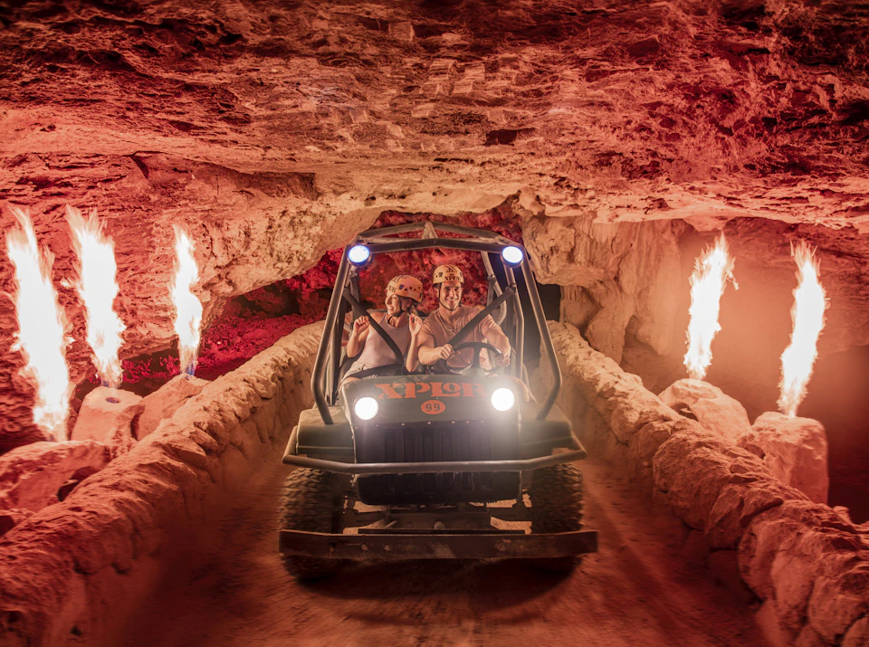 Two people in helmets driving an all-terrain vehicle through a cave tunnel with flames on both sides.