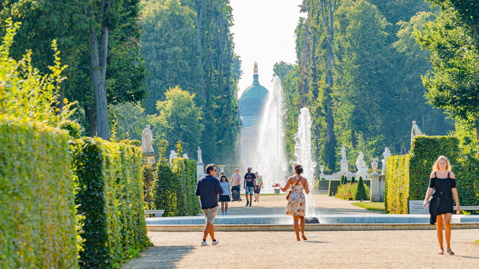 Persones caminant per un camí enjardinat amb fonts i estàtues en un jardí, envoltades d&#39;arbres alts i setos retallats.