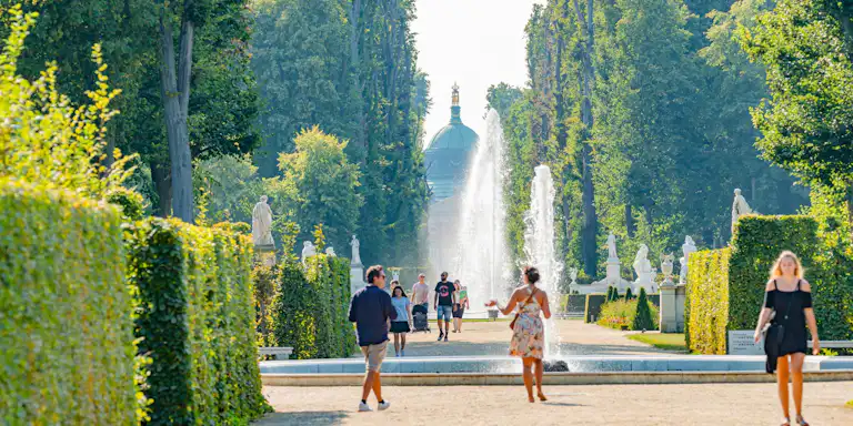 Menschen, die einen landschaftlich gestalteten Weg mit Springbrunnen und Statuen in einem Garten entlanggehen, umgeben von hohen Bäumen und gestutzten Hecken.