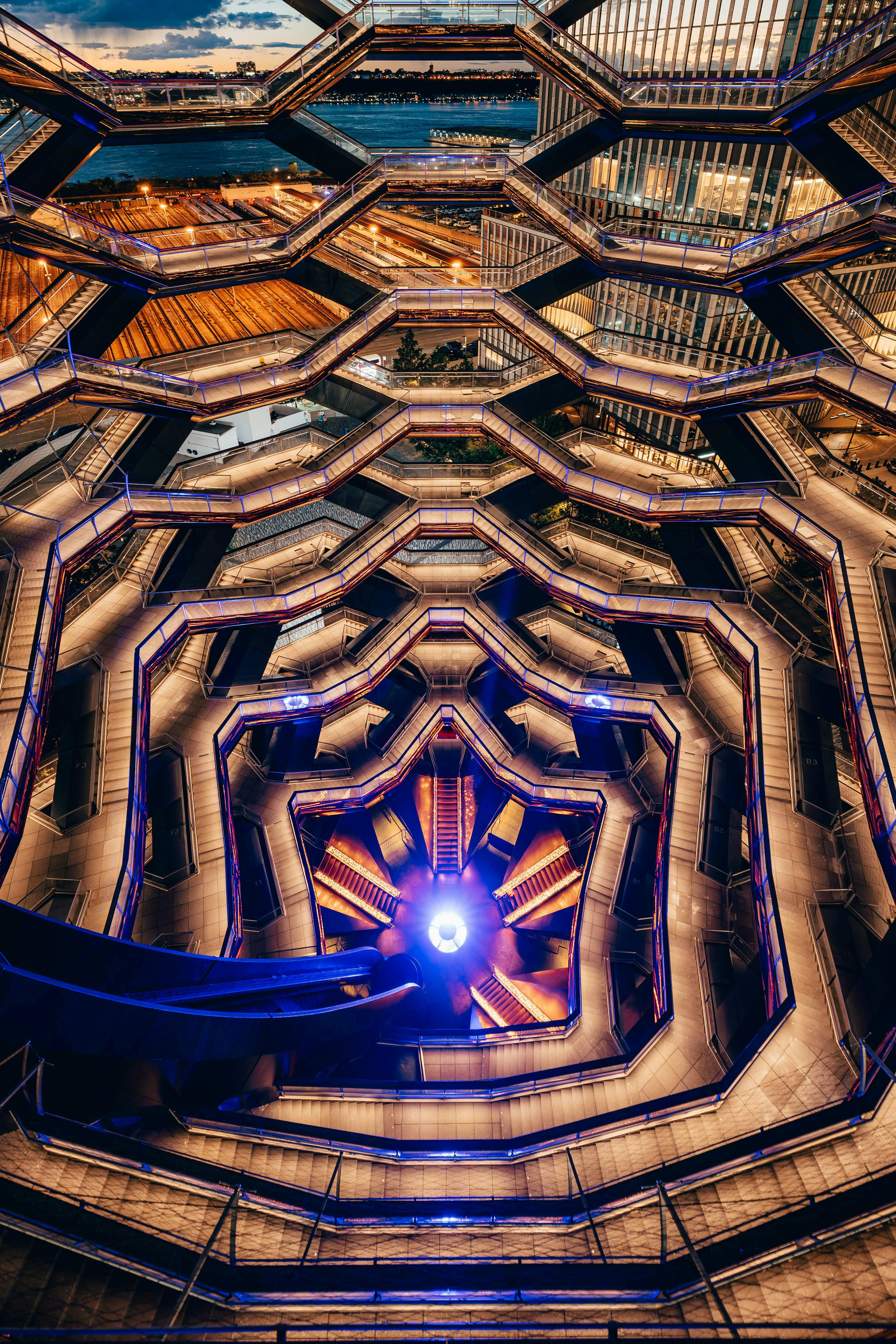 Geometric structure with multiple hexagonal levels, illuminated by purple and yellow lights, seen from an upward perspective.