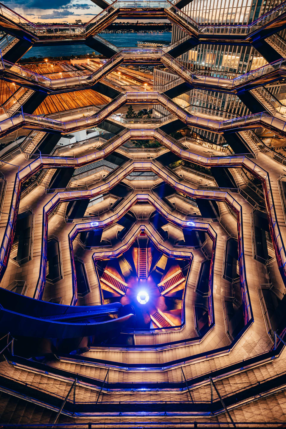 Geometric structure with multiple hexagonal levels, illuminated by purple and yellow lights, seen from an upward perspective.