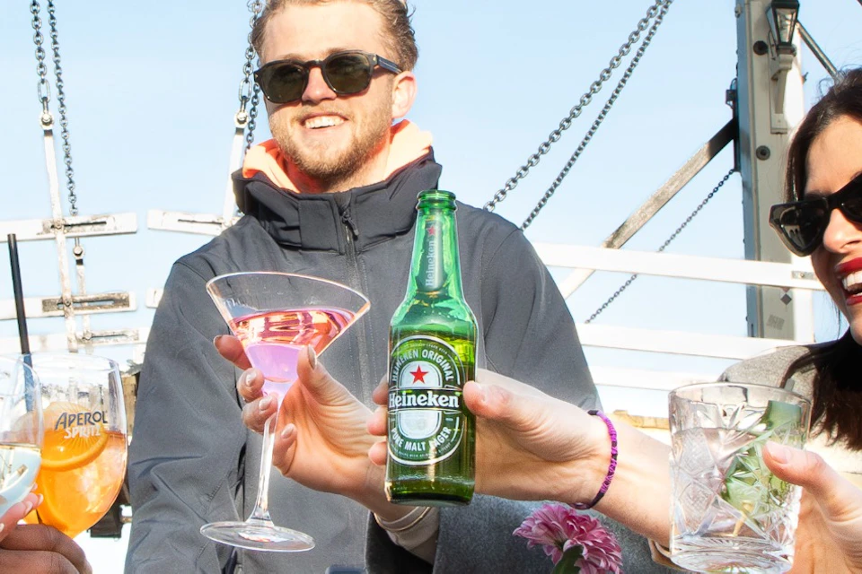 A group of people enjoying drinks and smiling under a clear blue sky, with a bridge structure visible in the background.