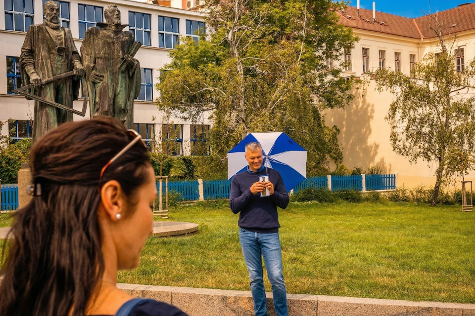 A person holding a blue and white umbrella stands on grass near buildings, with another person partially visible in the foreground.