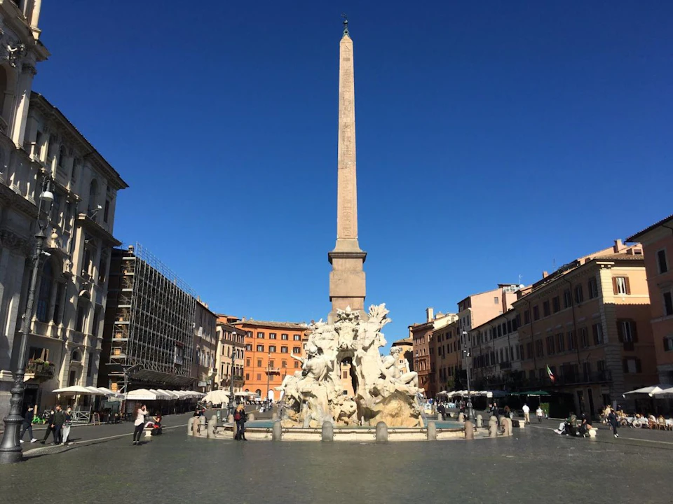 A tall obelisk stands in the center of a plaza fountain, surrounded by historic buildings under a clear blue sky. People are walking around.