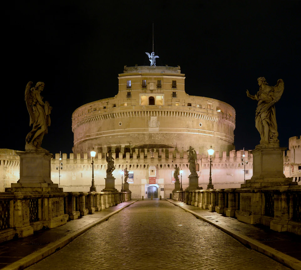 A large round fortress with a statue on top, flanked by angel statues and street lamps on a cobblestone path at night.