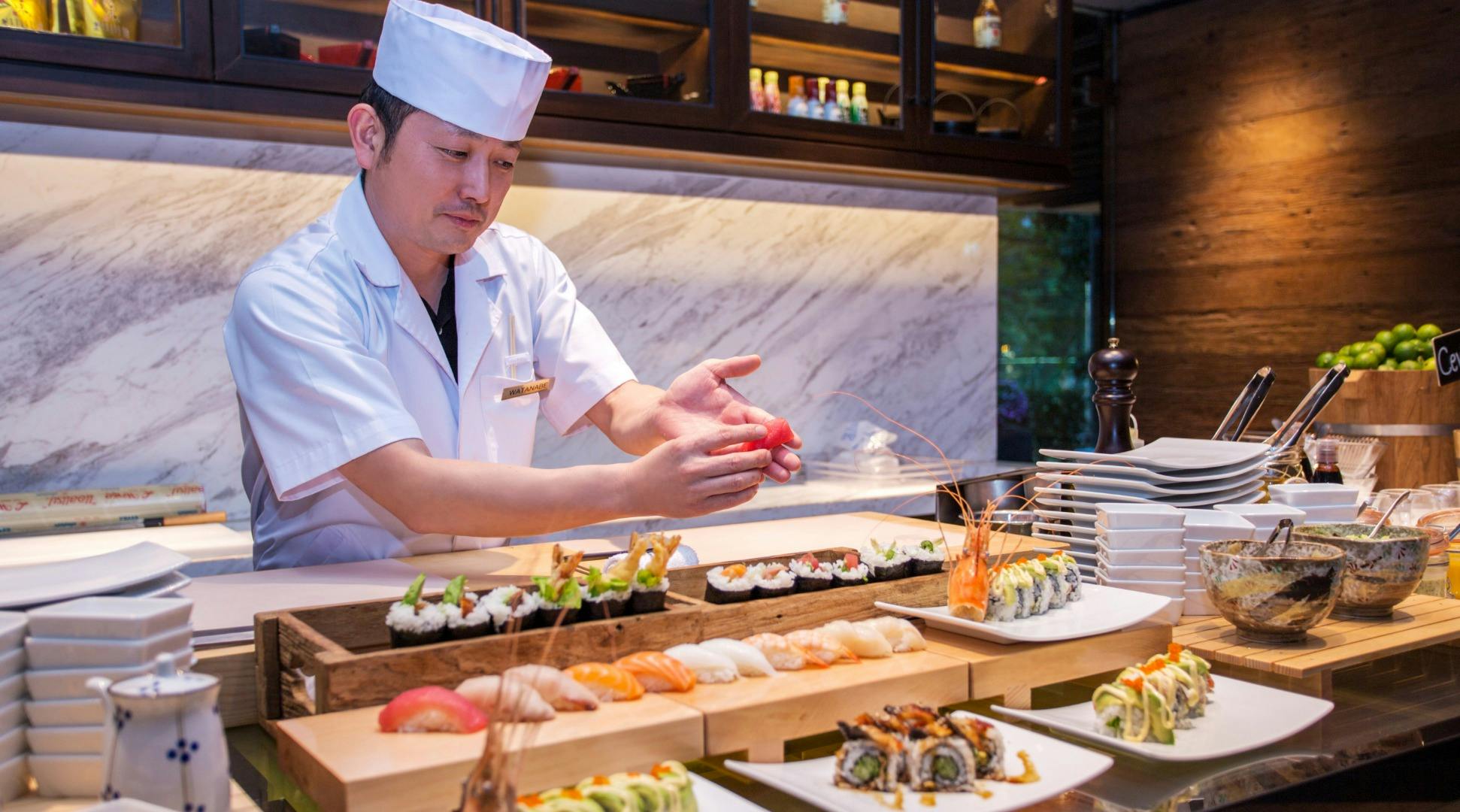 A sushi chef in a white uniform preparing sushi behind a counter with various sushi items displayed on wooden trays.