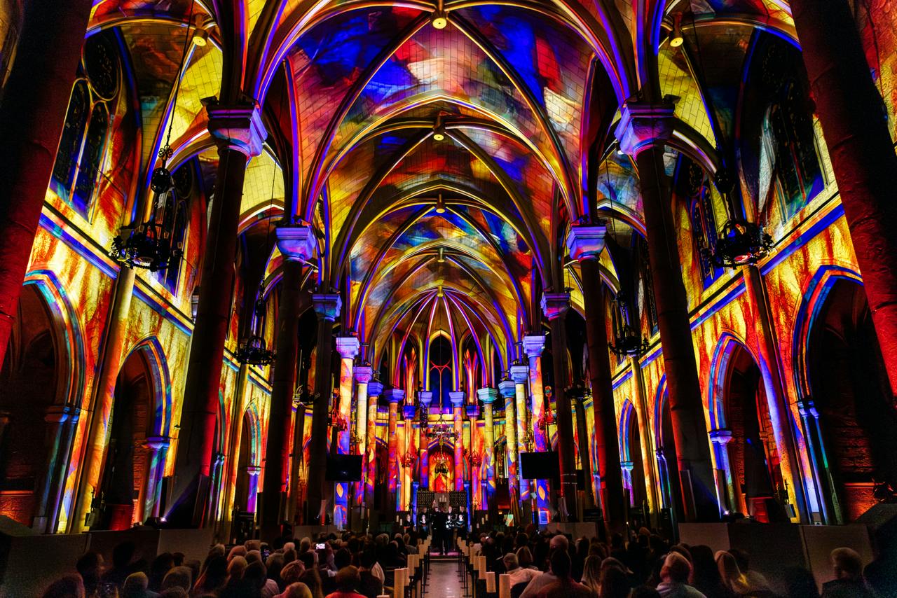 Un intérieur de cathédrale avec des projections lumineuses vibrantes et colorées sur les arcs et les murs, rempli de personnes assises face à l'autel.