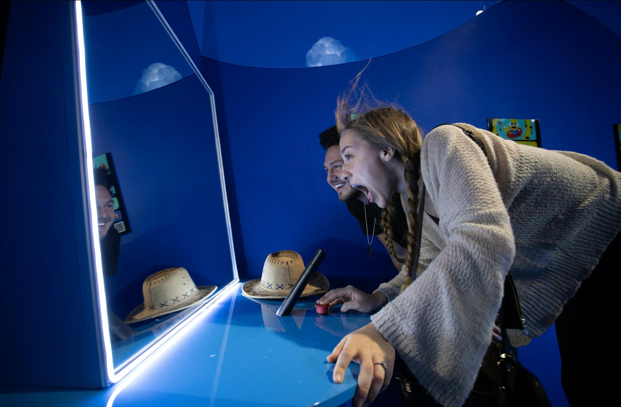 Two people interacting with a blue exhibit featuring a cowboy hat and mirrors, appearing excited and engaged.