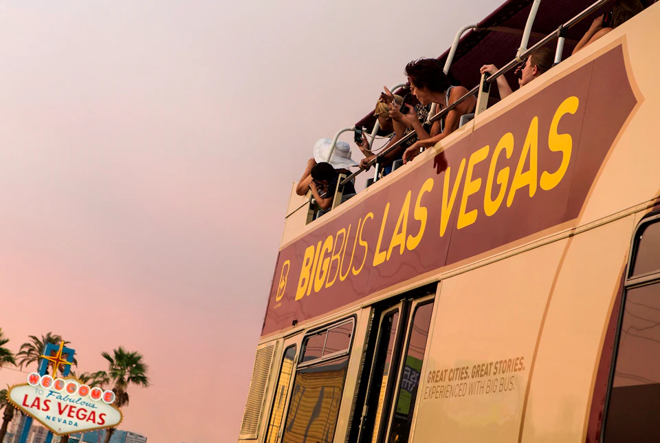 People on the upper deck of a "Big Bus Las Vegas" tour bus, taking photos. Palm trees and a pastel sky are in the background.