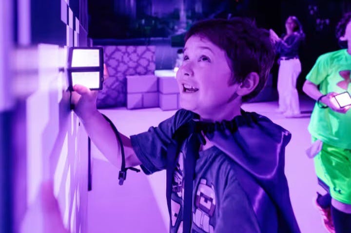 Ребенок в темном плаще улыбается, держа в руках освещенное устройство в комнате с фиолетовым освещением. Другие люди на заднем плане.
