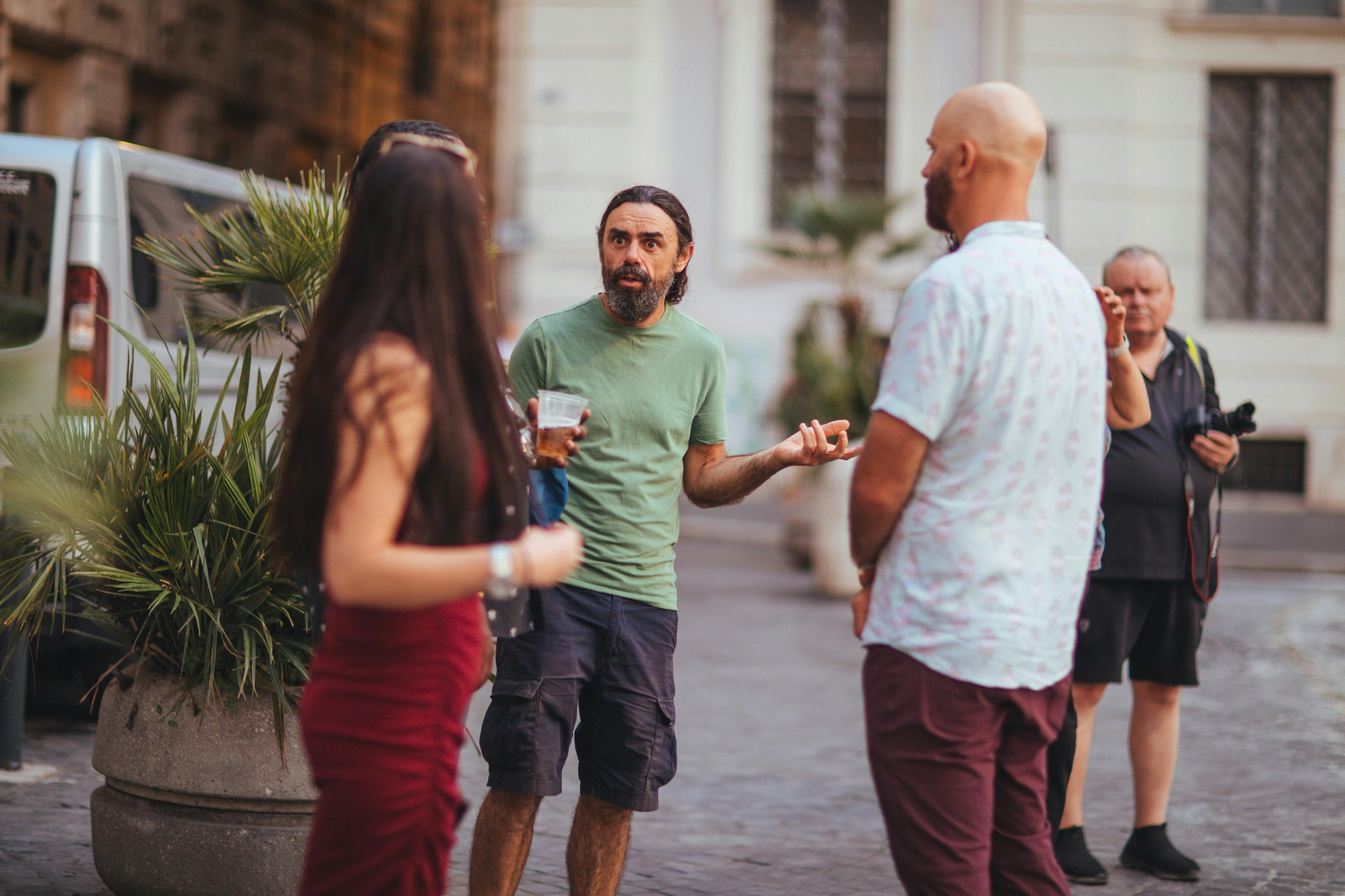 A group of people standing and talking outdoors in a courtyard-like setting with plants and a building in the background.