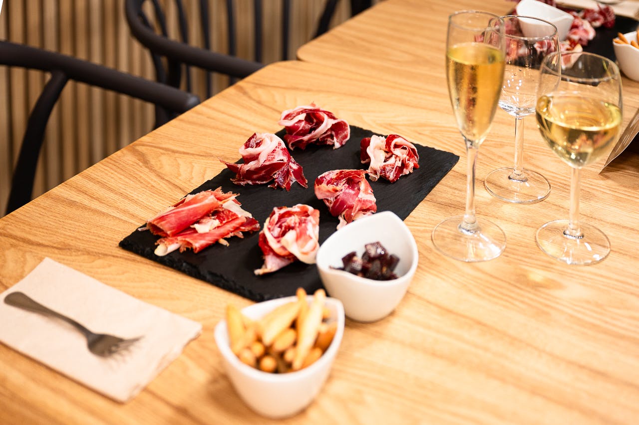A slate platter with cured ham, two small bowls with breadsticks and olives, and a glass of champagne on a wooden table.