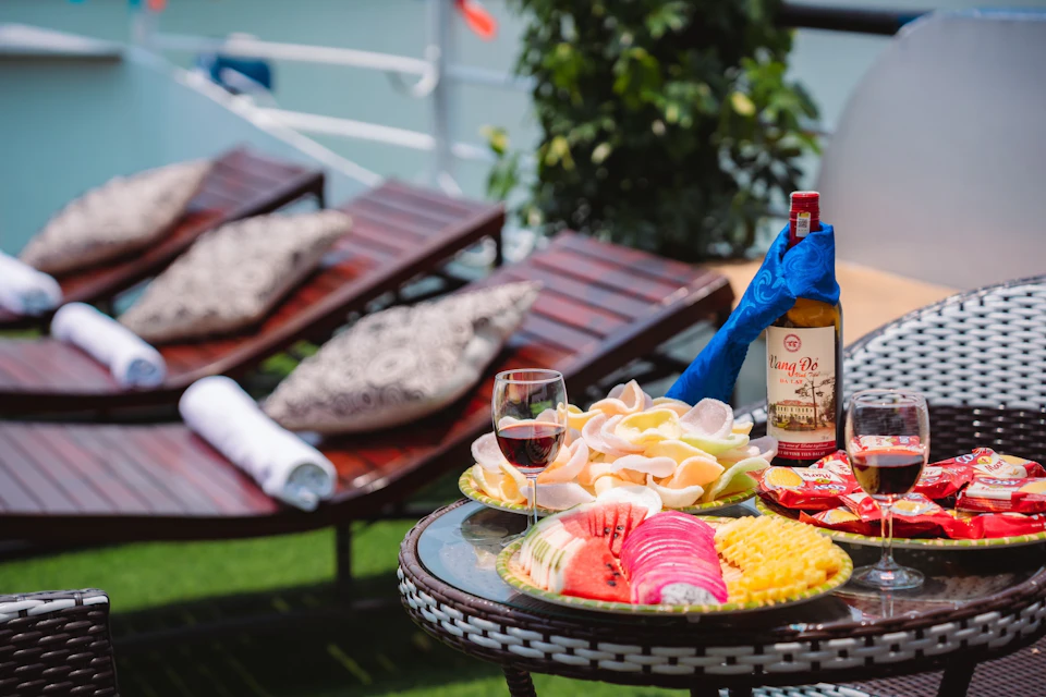 A table with fruit, wine, and snacks next to lounge chairs with pillows and a towel outside.