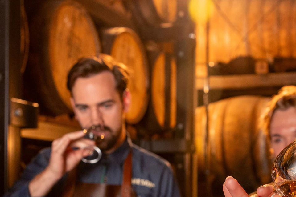 A group of people tasting drinks in a wooden barrel storage room, with glasses and a menu on the table.