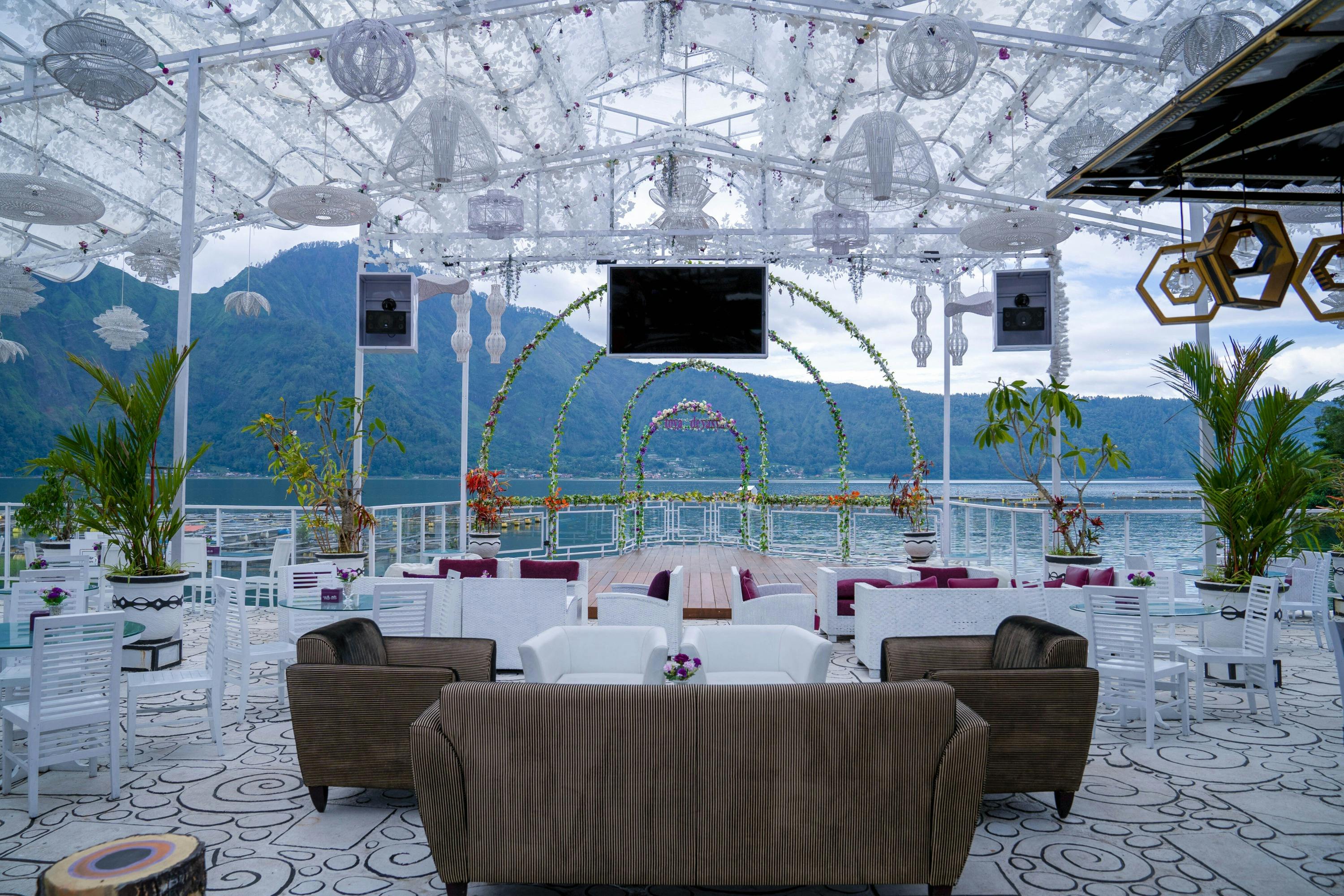 Un coin salon en plein air avec du mobilier blanc, des arches décoratives et une vue sur un lac entouré de collines verdoyantes.