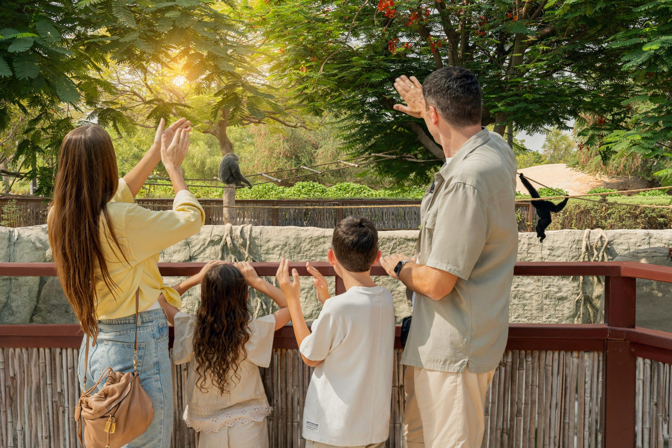 A family of four waves at a zoo exhibit, with two monkeys on a wooden structure under leafy trees.
