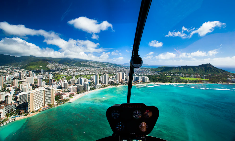 Aerial view from a helicopter cockpit overlooking a coastal city with high-rise buildings, beaches, and clear blue ocean under a bright sky.