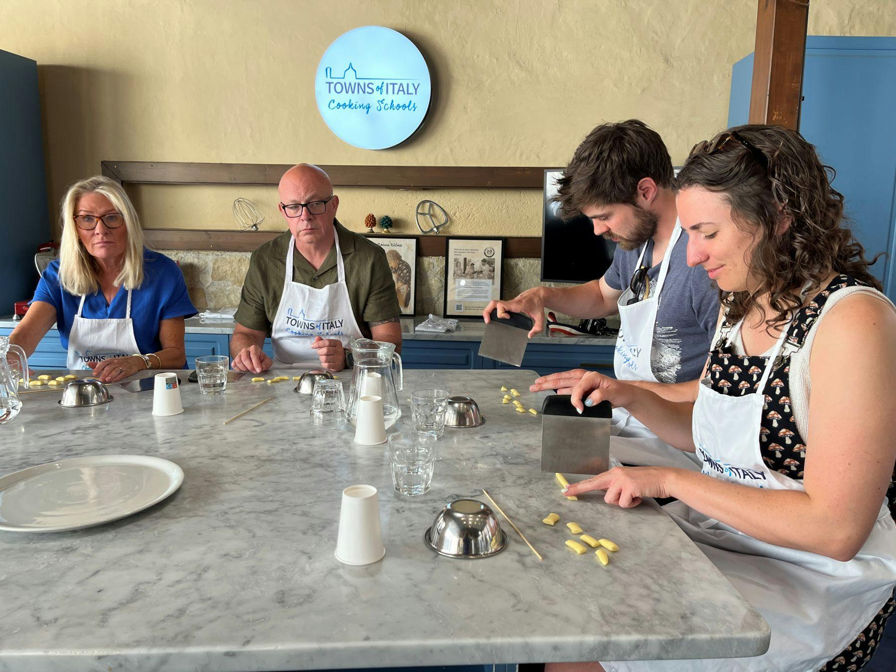 Four people participate in a cooking class, cutting small pieces of dough on a marble countertop. A logo sign is on the wall.