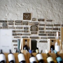 Vinflaskor på hyllor i förgrunden, med en stenvägg som visar många metallplattor i bakgrunden.