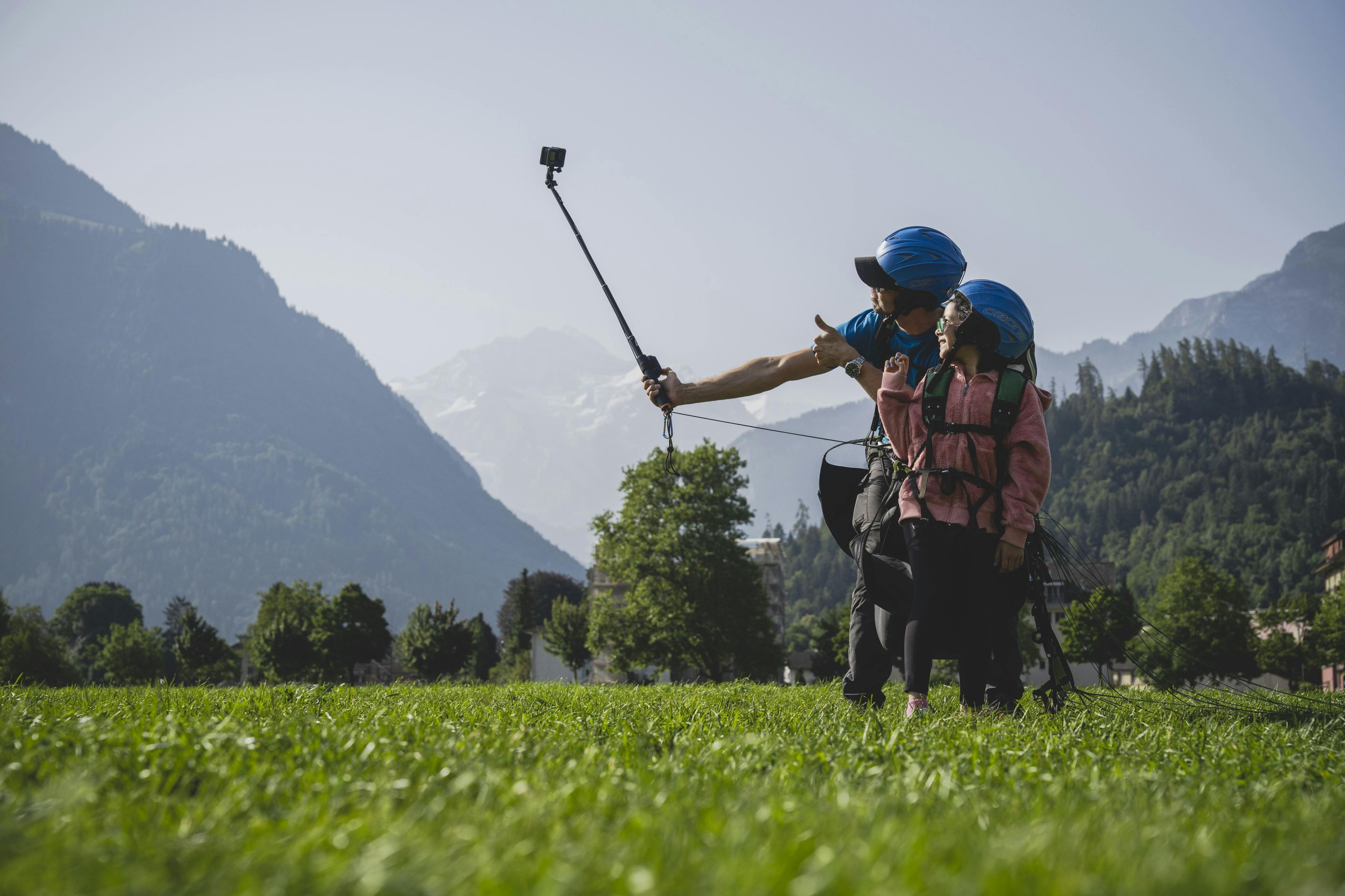 Deux personnes portant des casques, se préparant à faire du parapente dans un champ herbeux avec des montagnes en arrière-plan, prenant un selfie.