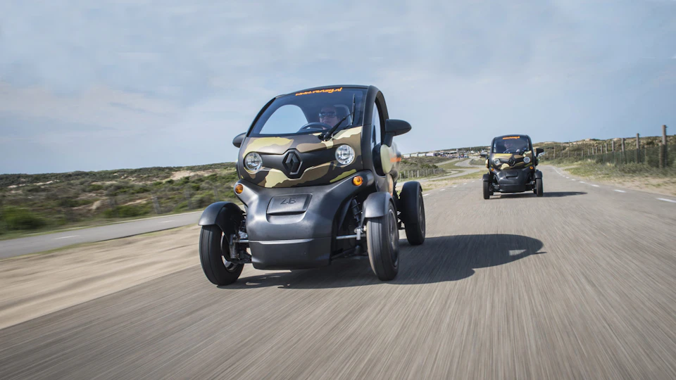Two small, camouflaged electric vehicles driving on a road with a landscape in the background.