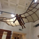 A wooden mannequin with wings modeled after Da Vinci's flying machine hangs suspended in a museum setting.