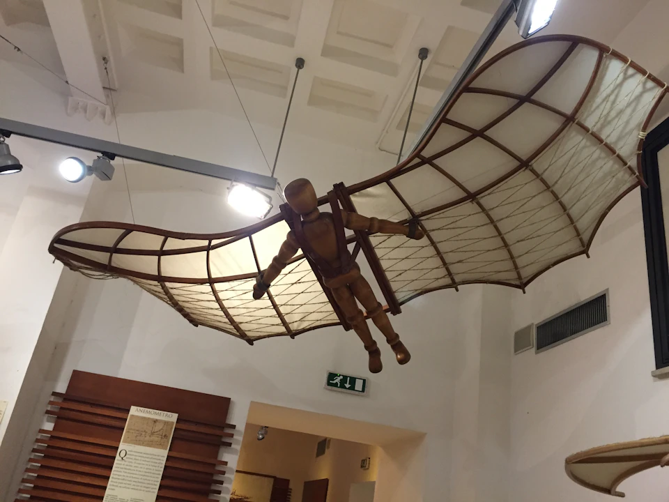 A wooden mannequin with wings modeled after Da Vinci's flying machine hangs suspended in a museum setting.