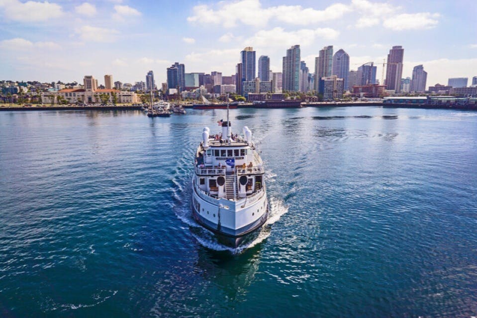 Ett litet kryssningsfartyg seglar mot kameran med en stadssilhuett i bakgrunden under delvis molnig himmel.