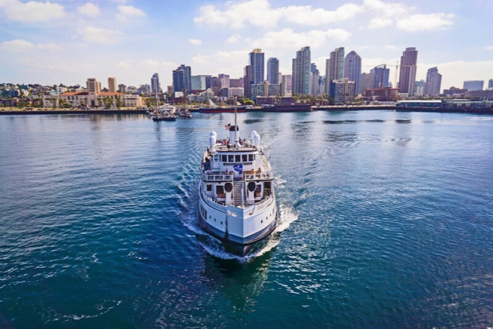 A small cruise ship sails towards the camera with a city skyline in the background under partly cloudy skies.