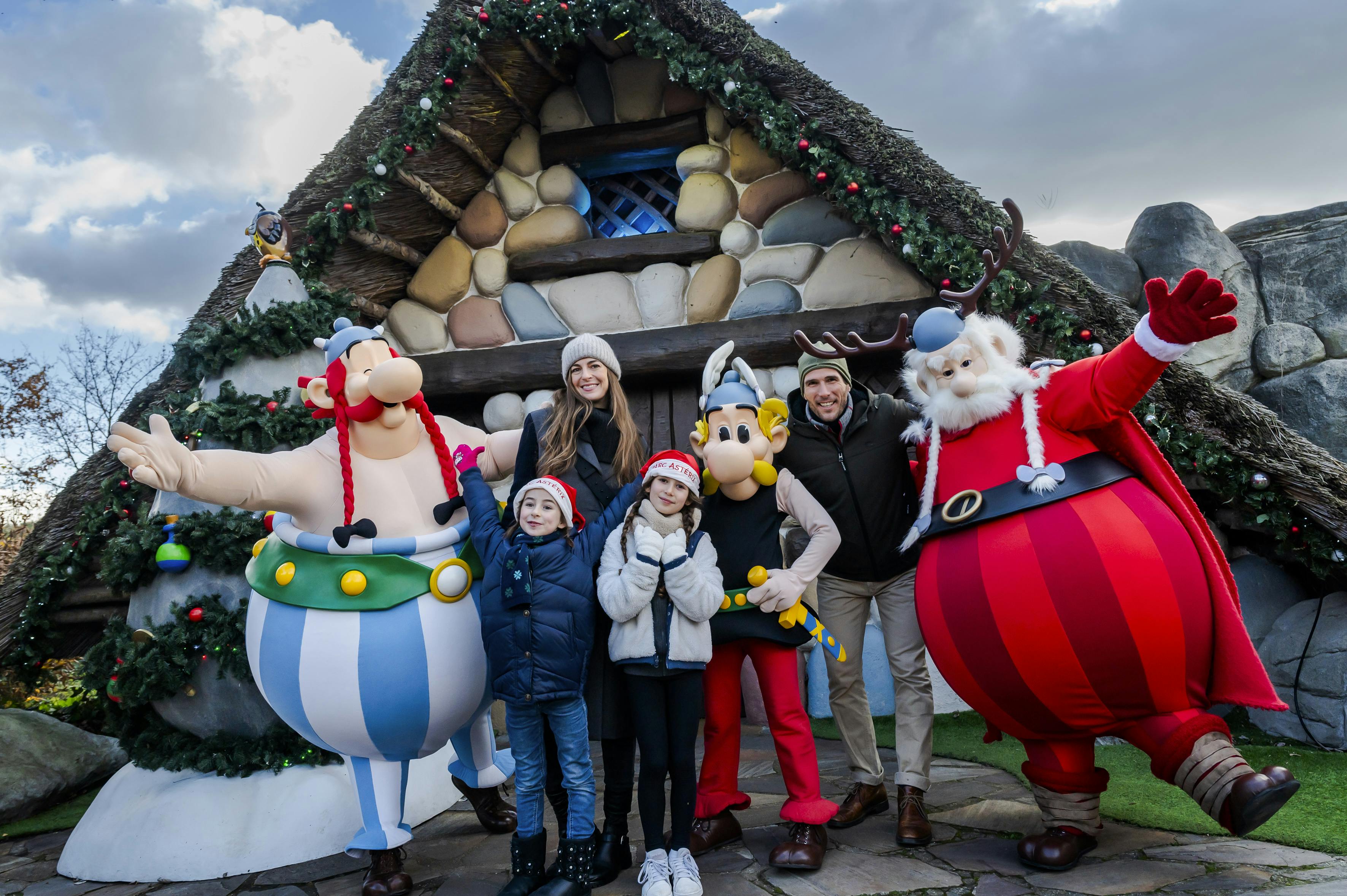 A family posing with cartoon characters in front of a festive stone house decorated with greenery and holiday lights.